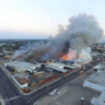 A large industrial fire with thick smoke rising into the sky and firefighters responding