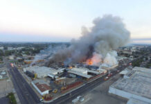 A large industrial fire with thick smoke rising into the sky and firefighters responding