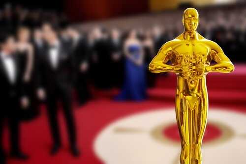 Golden Oscar statue in the foreground with blurred celebrities in formal attire in the background