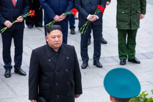 A political figure in formal attire standing solemnly during a ceremony with attendees holding flowers