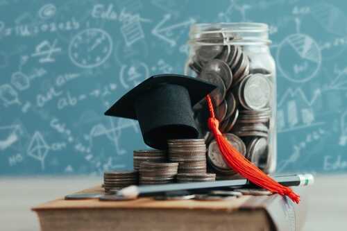 Graduation cap on a stack of coins next to a jar of coins and a pencil