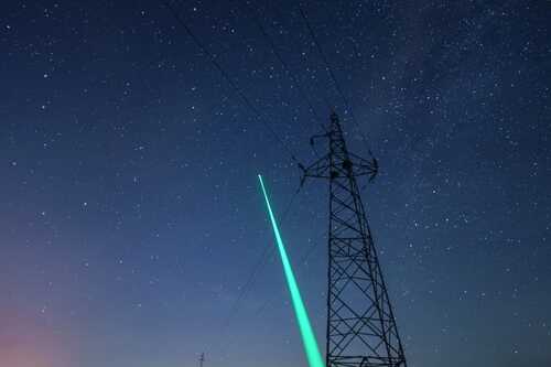 A green laser beam pointing towards a starry night sky near power lines