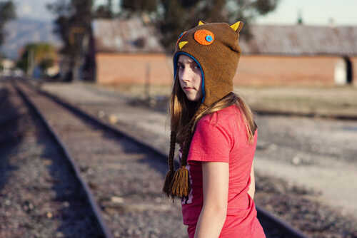 A girl wearing a playful brown hat stands on railroad tracks