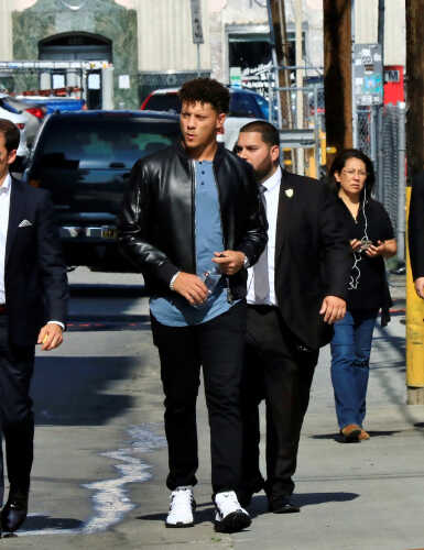 A group of men walking in an urban area, with one in a leather jacket and security personnel nearby