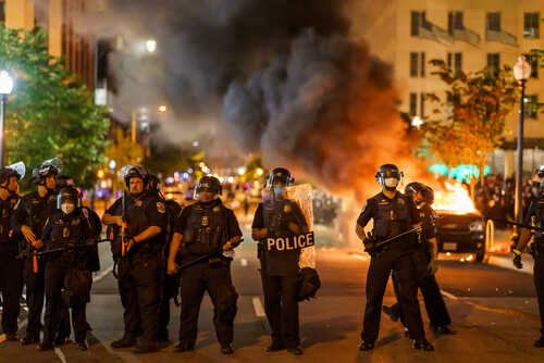 Police officers in riot gear facing a protest with smoke and fire in the background