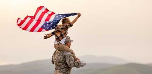 A child holding an American flag while sitting on a soldier's shoulders