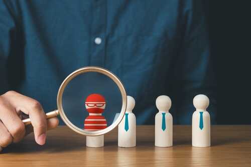 A hand holding a magnifying glass over a red and white striped figure among wooden figures