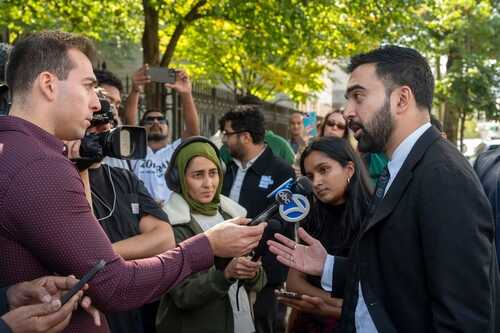 A man in a suit speaking to a reporter while surrounded by a crowd