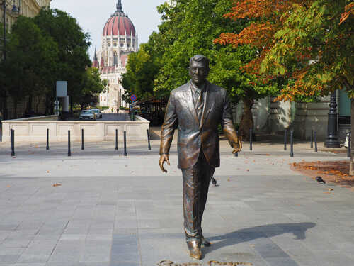 shutterstock_1817093903.jpg Bronze statue of a man in a suit walking through a city park with a historic building in the background