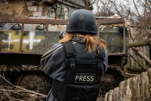 Journalist in a press vest aiming a camera towards a military vehicle