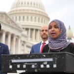 A woman in a hijab speaking at a podium outside the Capitol building, flanked by two men