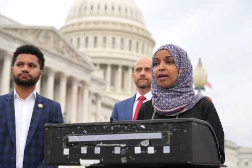 shutterstock_2279665109.jpg A woman in a hijab speaking at a podium outside the Capitol building, flanked by two men