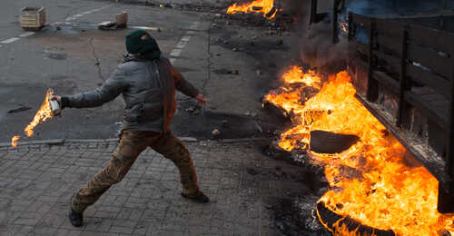 A person throwing a Molotov cocktail at a burning vehicle during a protest