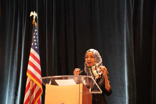 A woman in a hijab speaking passionately at a podium with an American flag in the background