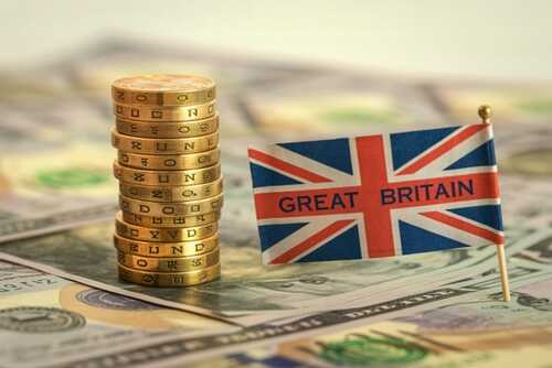 Stack of British pound coins next to a small flag of Great Britain on a background of dollar bills