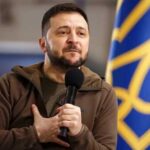 A political leader speaking passionately into a microphone with a national flag in the background