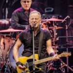 A guitarist performing on stage with a drummer in the background