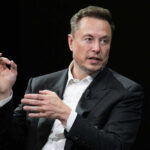 Man in suit speaking, black background, gesturing with hands.