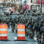A line of soldiers in military uniforms marching past traffic barriers in an urban setting