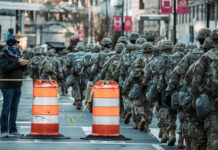 A line of soldiers in military uniforms marching past traffic barriers in an urban setting