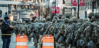 A line of soldiers in military uniforms marching past traffic barriers in an urban setting