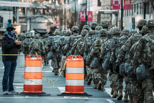 A line of soldiers in military uniforms marching past traffic barriers in an urban setting