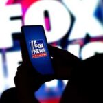 Fox Poll BOMBS Trump Iran Push A person holding a smartphone displaying the Fox News logo against a colorful background
