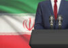 Khamenei Hidden in Secret Bunker! A suited individual standing behind a podium with microphones in front of the Iranian flag