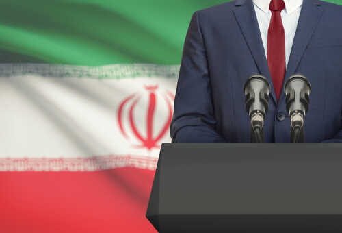 Businessman,Or,Politician,Making,Speech,From,Behind,The,Pulpit,With A suited individual standing behind a podium with microphones in front of the Iranian flag
