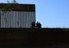 El Mencho Down — Mexico Explodes Silhouetted figures by tall fence against blue sky