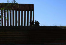 El Mencho Down — Mexico Explodes Silhouetted figures by tall fence against blue sky