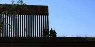 Silhouetted figures by tall fence against blue sky