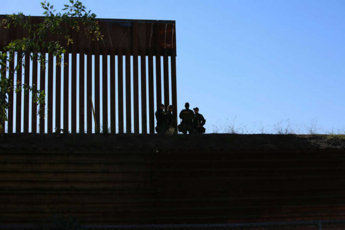 Silhouetted figures by tall fence against blue sky