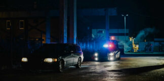 A police car with flashing lights parked next to a dark vehicle in a dimly lit urban area