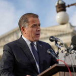 Man speaking at a podium with microphones outside
