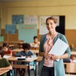 A smiling teacher holding papers in a classroom with students working at their desks