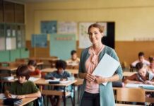 School Indoctrination Bombshell Hits K-12 A smiling teacher holding papers in a classroom with students working at their desks