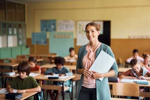 Happy,Teacher,During,A,Class,At,Elementary,School,Looking,At A smiling teacher holding papers in a classroom with students working at their desks
