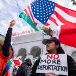 Two women at a protest holding flags and a sign against deportation