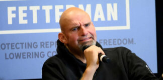 Man holding microphone, sitting under Fetterman sign.