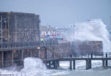 A man in a yellow jacket walking along a pier during a storm with crashing waves