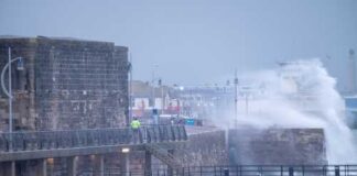A man in a yellow jacket walking along a pier during a storm with crashing waves