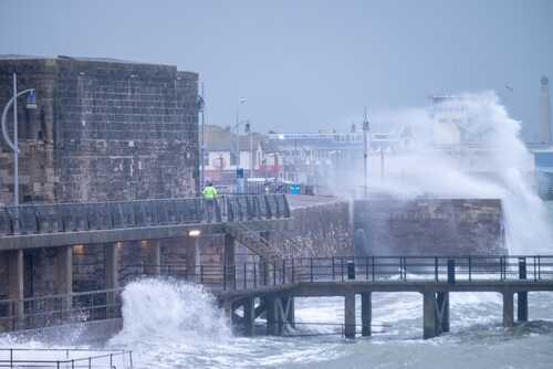 A man in a yellow jacket walking along a pier during a storm with crashing waves